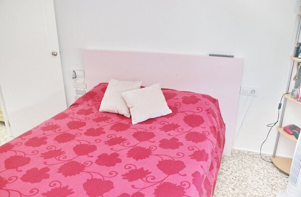 Un lit avec une couvre-lit à motifs floraux roses et deux oreillers, une tête de lit blanche et une petite étagère sur laquelle sont posés quelques objets.