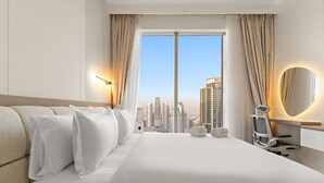 A hotel room with a large bed, a desk with a chair, a round mirror, and a view of a cityscape through a large window.