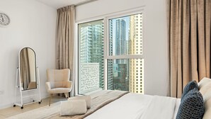 A modern hotel room with a large bed, a sitting chair, a standing mirror, and a view of tall buildings through large windows. - Stylish 1BR w/ JBR Beach Access, Dubai Marina & Ain Dubai Views (JBR)