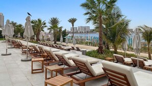A row of sun loungers with umbrellas, palm trees, and a view of a beach and buildings in the background. - Stylish 1BR w/ JBR Beach Access, Dubai Marina & Ain Dubai Views (JBR)
