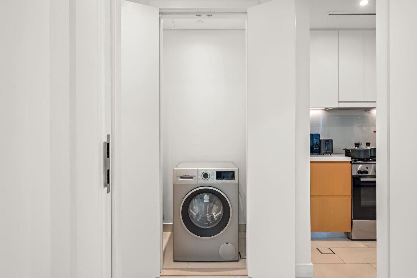 A compact laundry room with a front-loading washing machine, a small countertop, and a kitchen area visible through an open door.