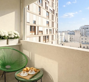 Un balcón con una silla verde, una pequeña mesa con cruasanes y café, y vista a un paisaje urbano.