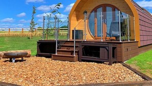 Uma cabana moderna de madeira curva, com fachada de vidro, um deck e um braseiro em primeiro plano.