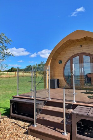A wooden cabin with a circular window, a wooden deck with metal railings, and a grassy field in the background.