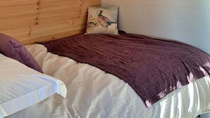 Ein Schlafzimmer mit schrägem Dach, einem Bett mit violetter Decke, einem Kissen mit einem Hahn-Motiv und einem an der Wand befestigten Regal mit Büchern und einem Bilderrahmen.
