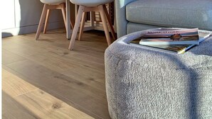 Ein gemütliches Zimmer im Dachgeschoss mit schrägen Holzbalken, einem Oberlicht, einem weißen Schreibtisch, zwei Stühlen, einem grauen Sofa und einer Zeitschrift auf einem Hocker.