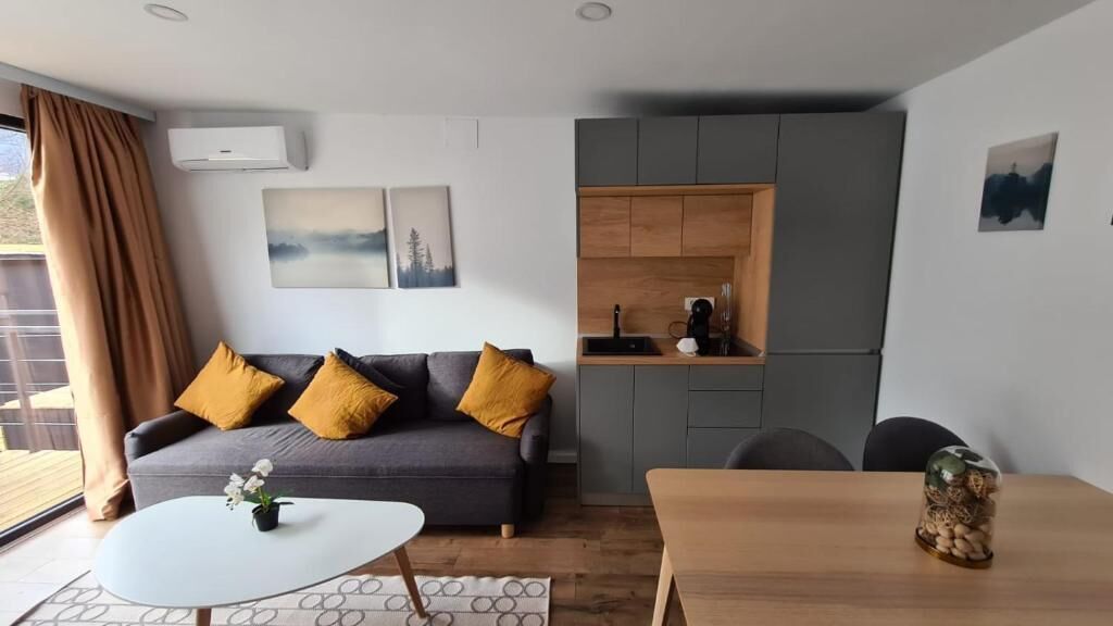 A modern living room with a grey sofa, a small round table, and a wooden dining table. There is a kitchen area with grey cabinets and a sink.