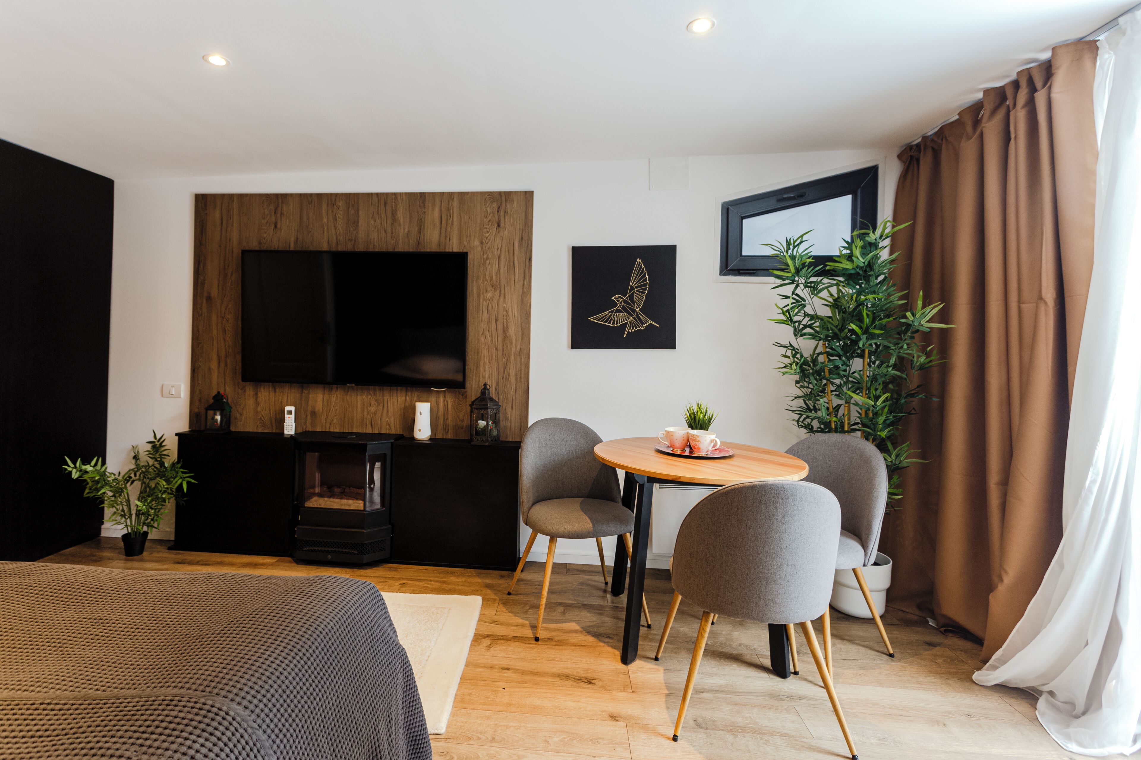 Ein modernes Hotelzimmer mit einer Wand in Holzoptik, einem Flachbildfernseher, einem runden Tisch mit Stühlen, einem Bett und einer Topfpflanze.