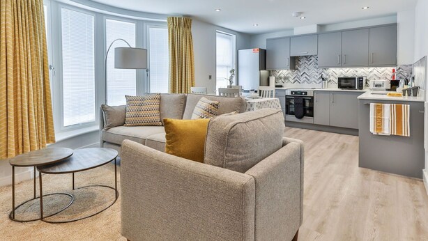 A modern living room with a sofa, a coffee table, and a kitchen area with grey cabinets and a herringbone backsplash.