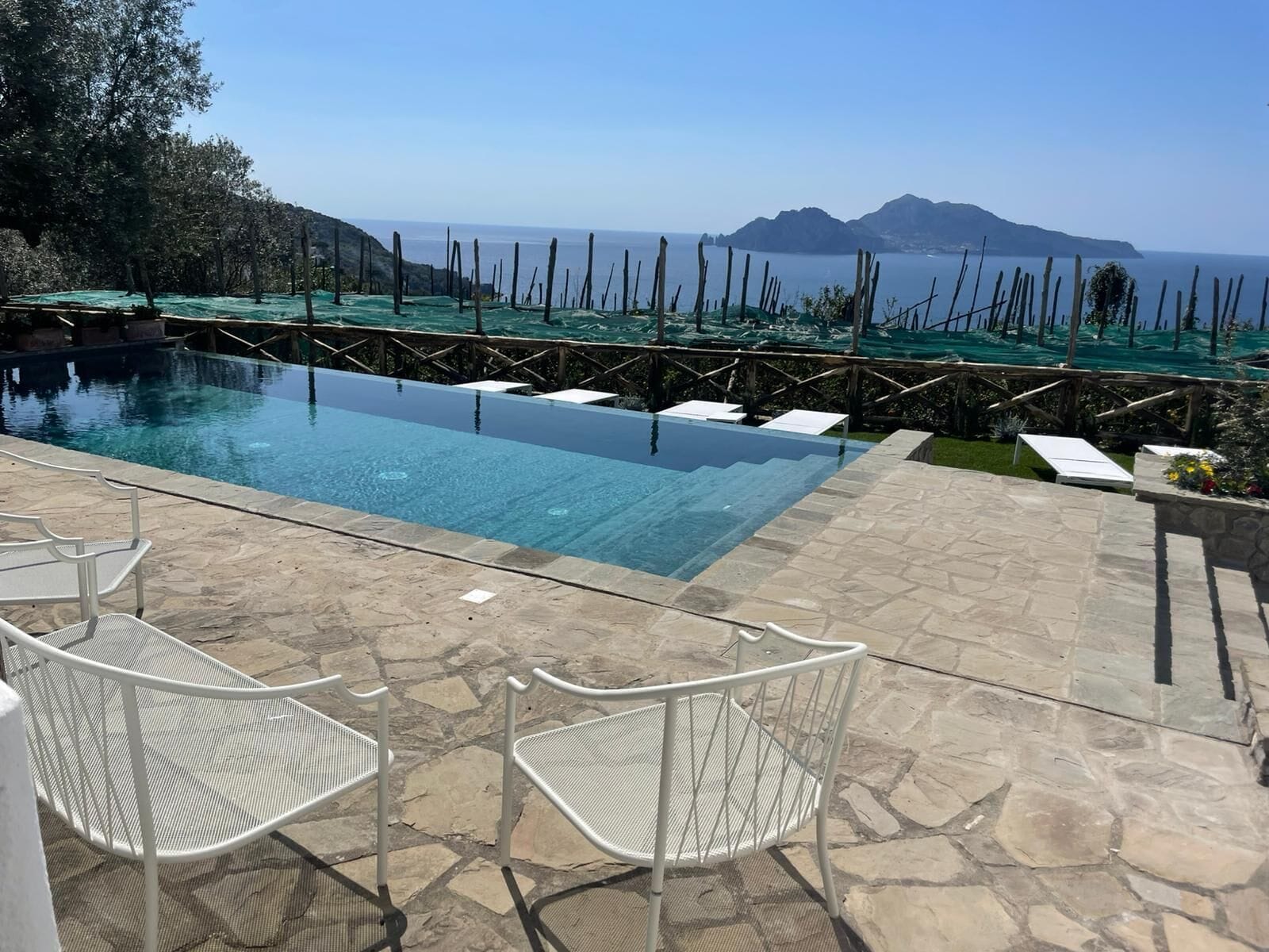 A clear swimming pool with a view of the sea and mountains, surrounded by stone flooring and white metal chairs.