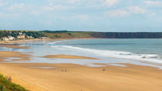 A coastal beach with a sandy shore, a few people, and buildings in the background.