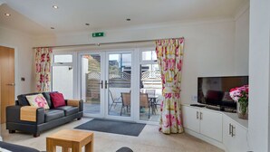A modern living room with a sofa, a coffee table, and a TV. There is a sliding glass door leading to an outdoor area with a table and chairs.