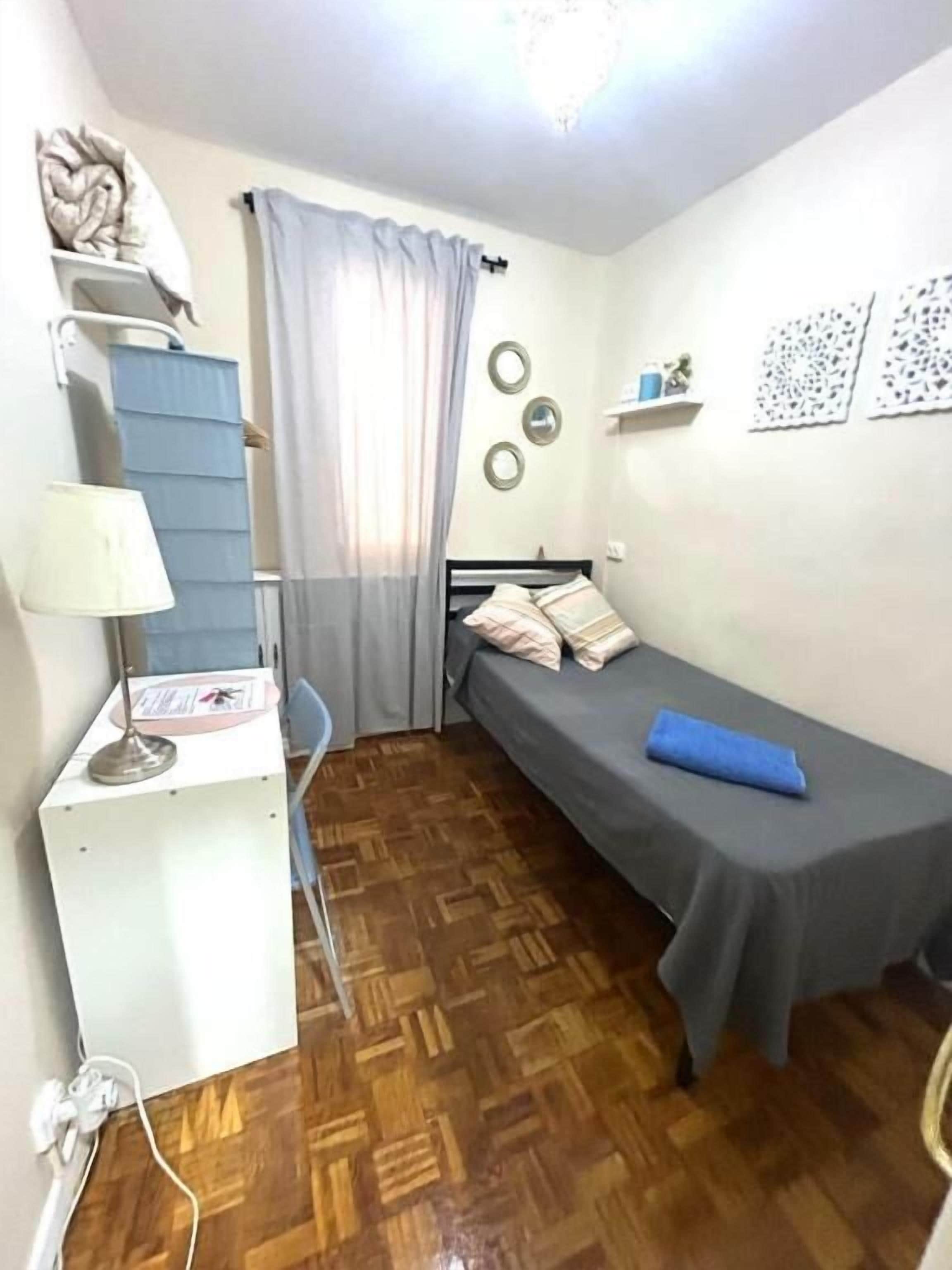 A small, compact room with a single bed, a desk, and a chair. There is a window with curtains, a wall-mounted shelf with decorative items, and a wooden floor.