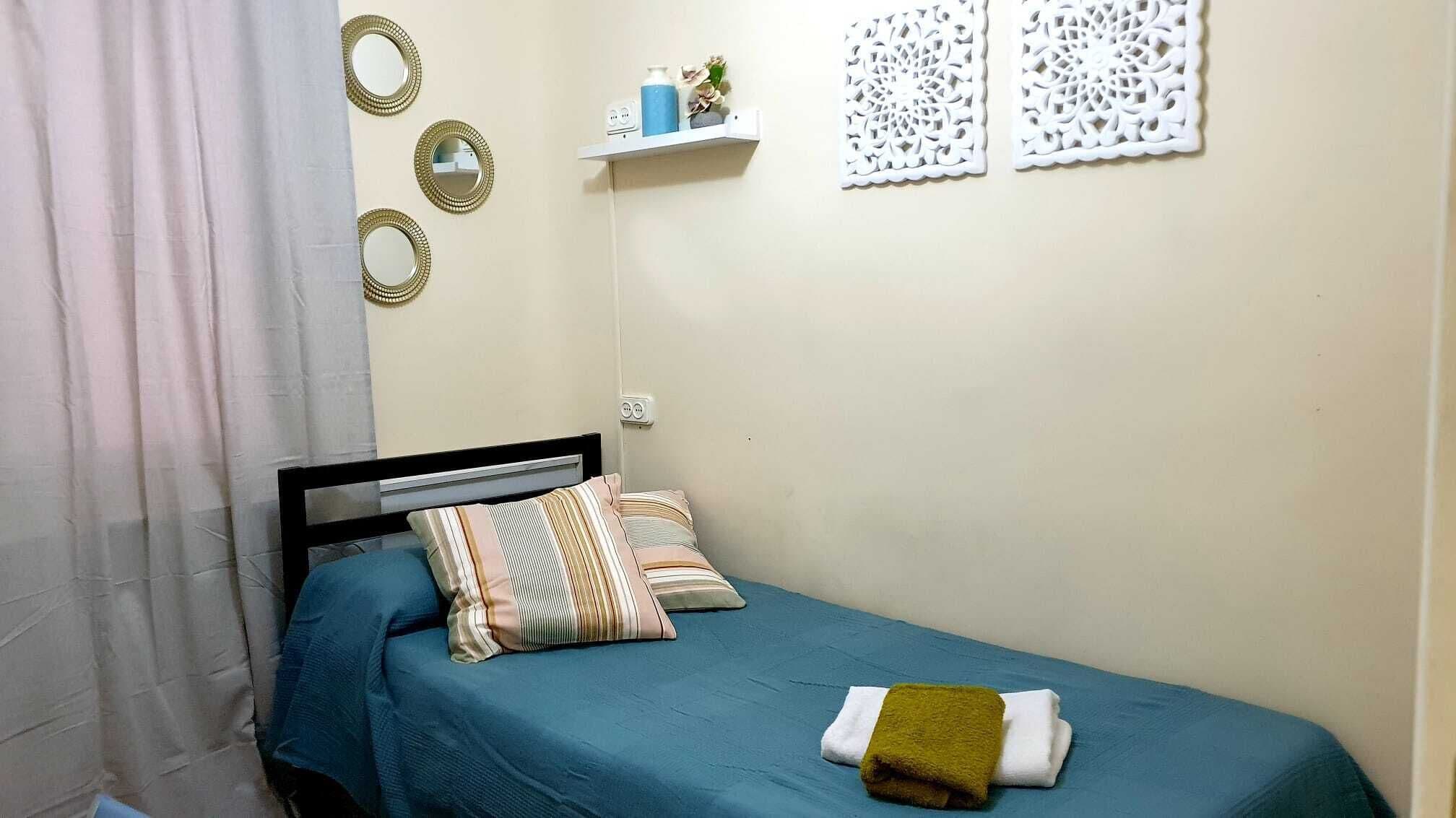 A small, neatly arranged bedroom with a single bed, a blue bedspread, and a few pillows. There are wall decorations and a shelf with items on it.