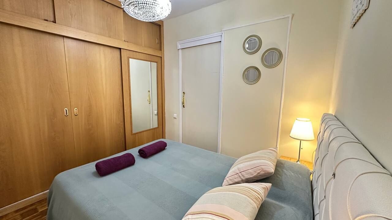A bedroom with a bed, a wardrobe, a mirror, a door, and a nightstand with a lamp.