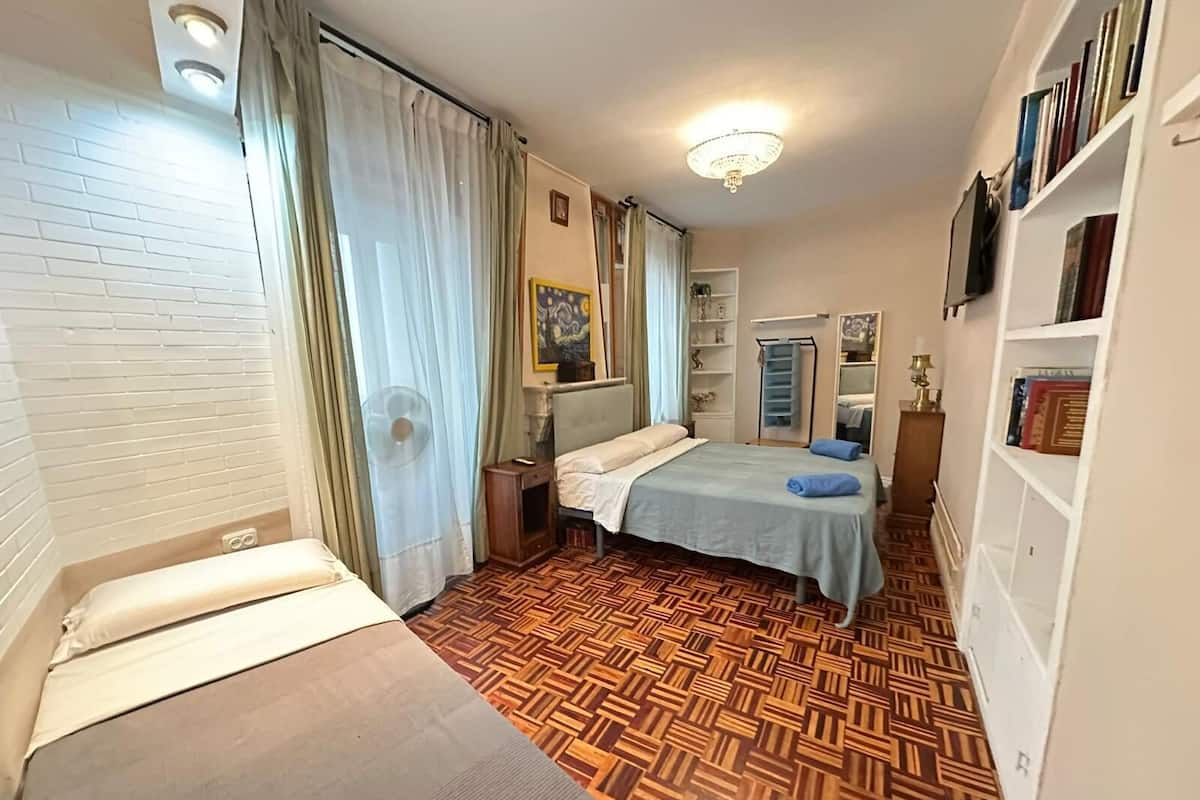 A hotel room with a bed, a small table, a fan, a mirror, a bookshelf, and a patterned wooden floor.