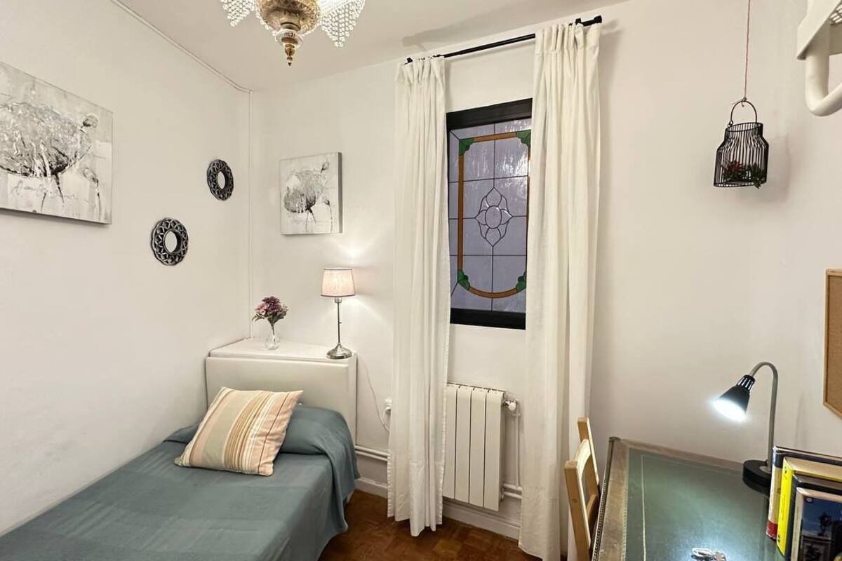 A bedroom with a bed, a nightstand, a lamp, a window with curtains, a radiator, and wall art.