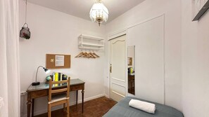 A bedroom with a bed, a desk with a chair, a hanging light fixture, a wardrobe, and a mirror.