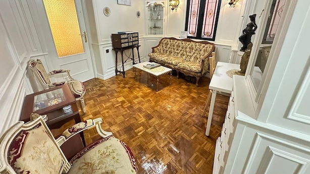 A spacious living room with a wooden parquet floor, a floral sofa, a small table, and a decorative cabinet. - Habitaciones en Luisa (Madrid)