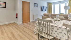 A modern dining area with a wooden floor, a dining table set with white tablecloths, and a sofa in the background. - Beautiful apartment for 4 guests with WIFI, TV and pets allowed (Filey)