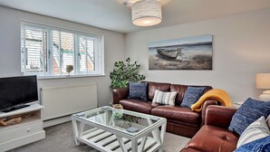 Un salon avec un canapé en cuir marron, une table basse en verre, un téléviseur posé sur un meuble blanc, une fenêtre avec des stores et un tableau représentant un bateau accroché au mur.