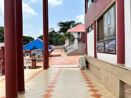 Área externa coberta com pilares vermelhos, piso de azulejos e um edifício com fachada vermelha e branca.