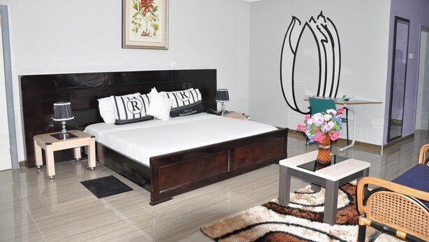 A modern bedroom with a large bed, bedside tables, a nightstand, a desk with a chair, a vase of flowers, and a framed picture on the wall. - Royal Lee's Hotel (Tutu)