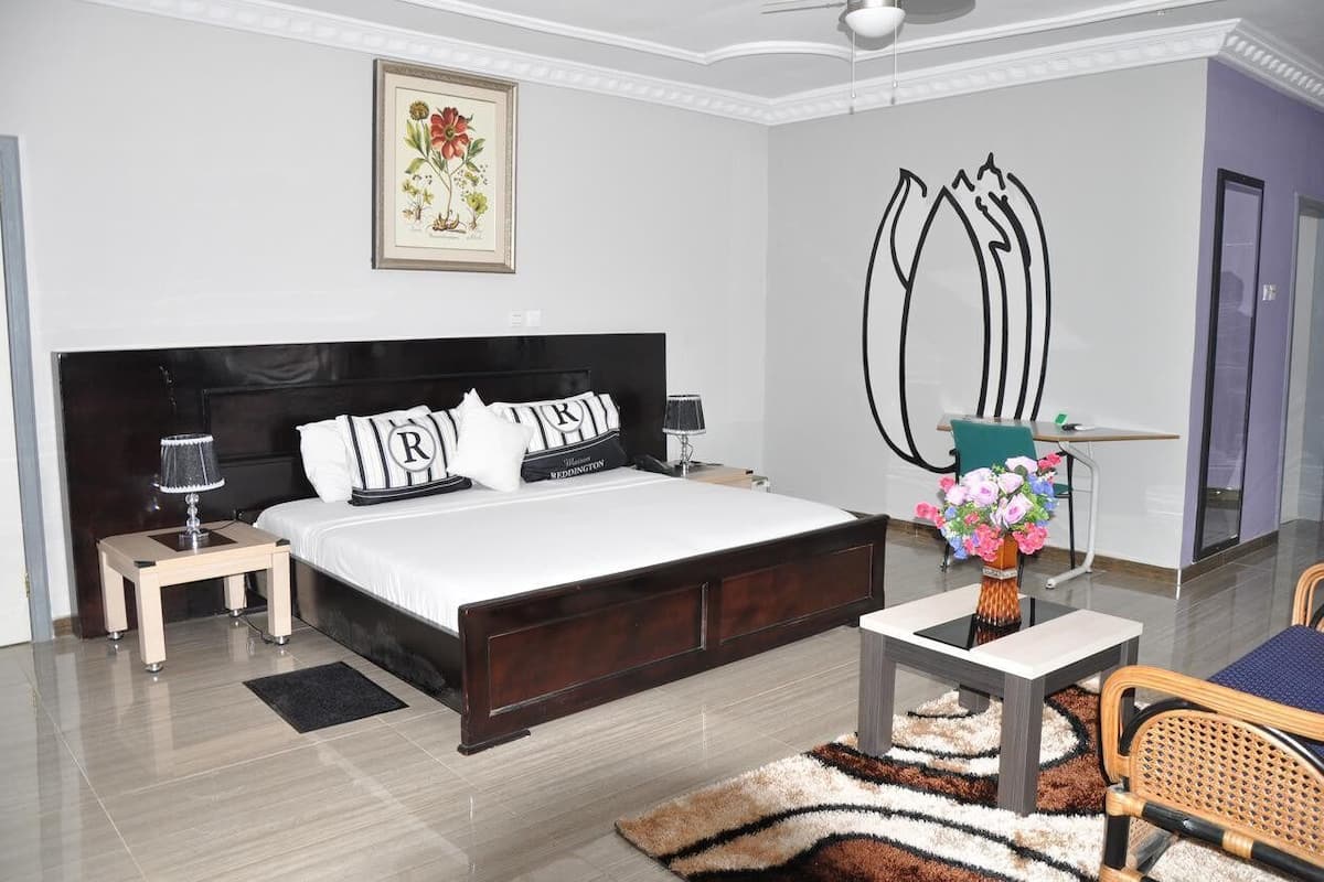 Un dormitorio moderno con una cama grande, mesitas de noche, un escritorio con silla, un jarrón de flores y un cuadro en la pared.