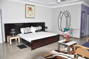 Quarto moderno com uma cama grande, mesas de cabeceira, um criado-mudo, uma escrivaninha com cadeira, um vaso de flores e um quadro na parede.