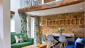 A cozy living room with a green sofa, a wooden coffee table, and a blue armchair. A stone wall and wooden beams are visible in the background.
