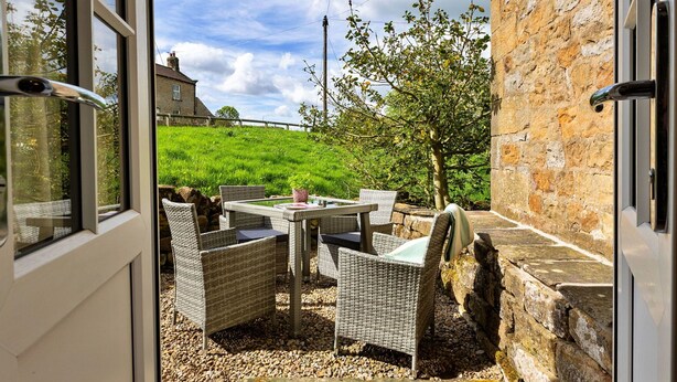 A patio with wicker furniture, a stone wall, and a view of a green field and house in the background.