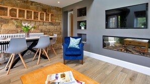 A modern living room with a stone wall, a fireplace, a blue armchair, and a dining area with a white table and chairs.