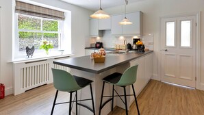 Cocina moderna con una isla central, dos taburetes de bar, luces colgantes y una ventana con vistas a zonas verdes.