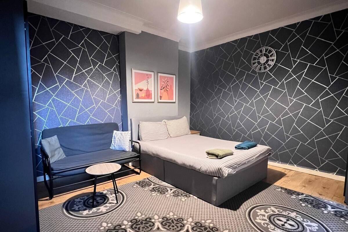 Un dormitorio moderno con una cama, un sofá y una mesita. La habitación presenta una pared con estampado geométrico y un tapete circular.