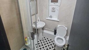 A bathroom with a glass shower enclosure, a sink, a toilet, and a window above the sink. - London E1 Hostel (London)