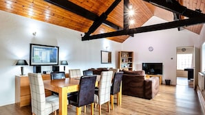Uma sala de estar ampla com teto de madeira, área de jantar com mesa e cadeiras de madeira, televisão e estante de livros.