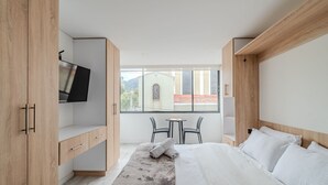 Ett hotellrum med en säng, en tv, ett skrivbord och ett fönster med utsikt över en byggnad och berg.