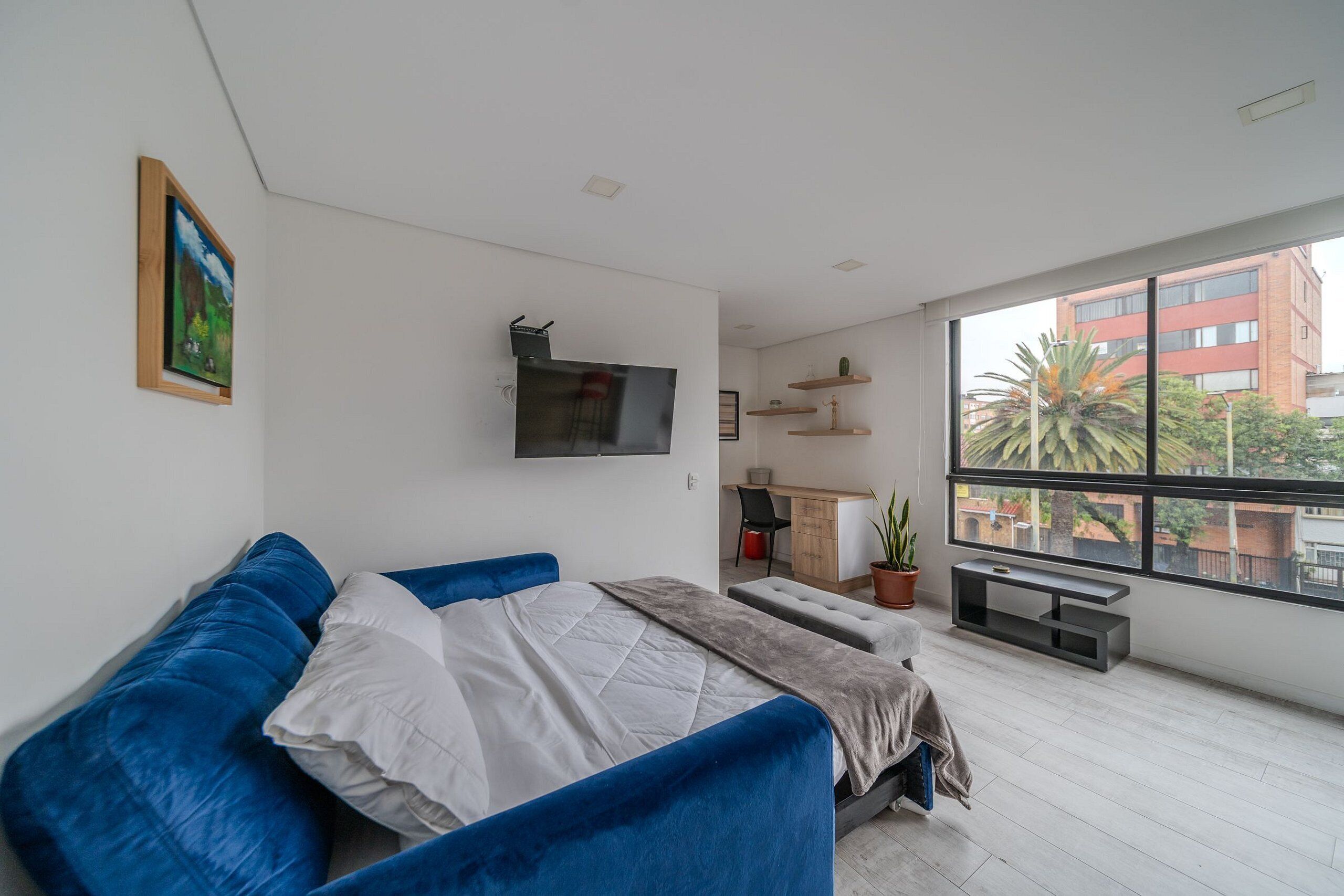 A modern bedroom with a large bed, a TV mounted on the wall, a desk with a chair, and a large window offering a view of buildings and trees.