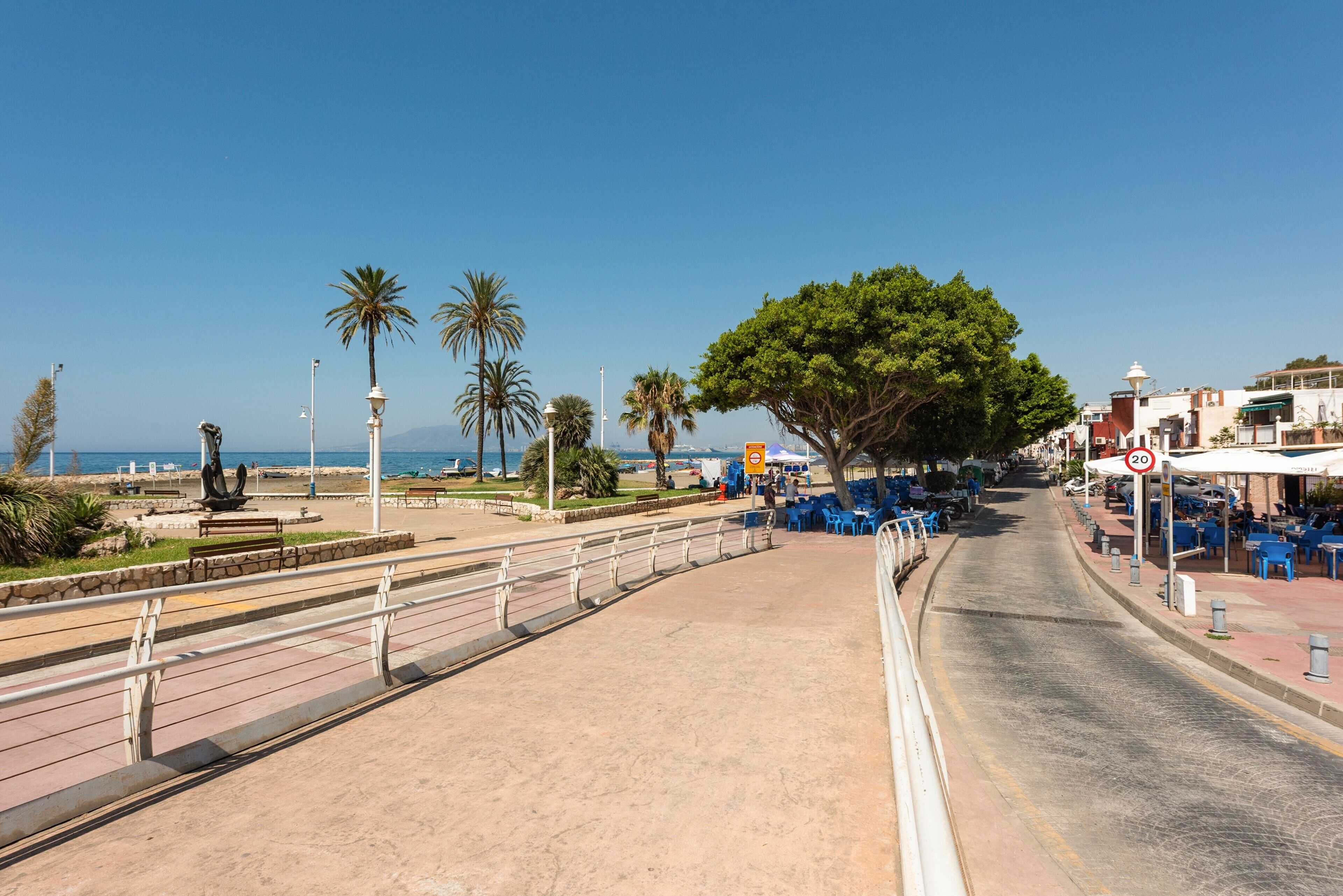 Eine Uferpromenade mit blauem Himmel, Palmen, einer Statue und Außenbestuhlung.