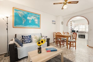 A living room with a sofa, a coffee table with a book and flowers, a dining area with chairs, a kitchen in the background, and a ceiling fan.