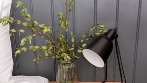 Un lit avec une literie blanche, une petite table de chevet en bois surmontée d’une lampe noire, un vase en verre contenant des feuilles vertes et un panier tressé avec un plaid à carreaux noirs et gris.