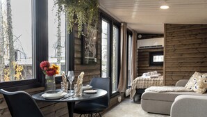 A modern interior with a dining table set for two, a sofa, and a television in a cozy room with wooden walls and large windows.