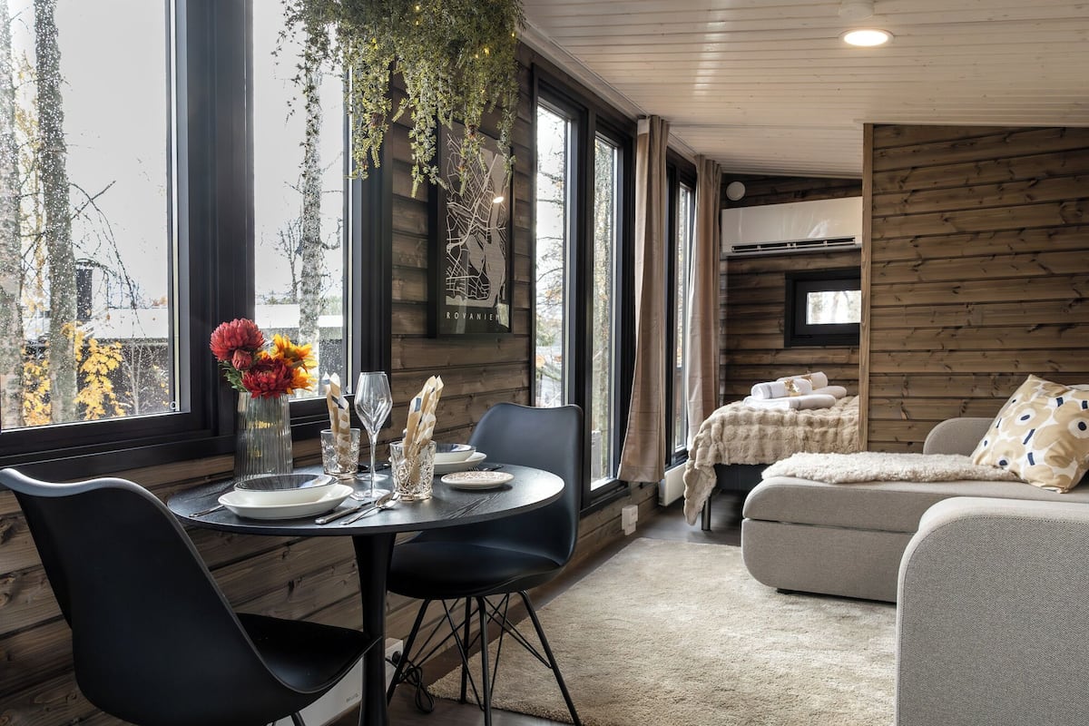 A modern interior with a dining table set for two, a sofa, and a television in a cozy room with wooden walls and large windows.