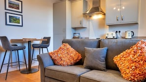 A modern living room with a grey sofa, two matching armchairs, and a wooden coffee table. There is a kitchen area with a stove, kettle, and toaster visible. - Nice apartment for 2 guests with WIFI and TV (Matlock)