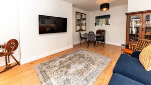 A living room with a TV, dining table, chairs, a blue sofa, a patterned rug, and two pendant lights.