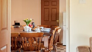 Een eetgedeelte met een houten tafel gedekt voor het ontbijt, inclusief brood, fruit en een koffiepot. Er staan stoelen rondom de tafel, en een houten deur is op de achtergrond te zien.
