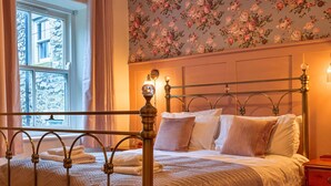 A neatly made bed with a metal frame, a floral wallpaper pattern on the wall, and a window with curtains.
