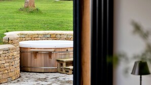 A stone hot tub with a wooden bench, surrounded by a stone wall and a grassy yard.