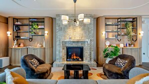 Una sala de estar moderna con chimenea, dos sillones, un sofá y estanterías empotradas con libros y plantas.