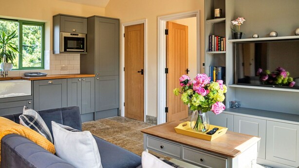 A modern living room with a grey sofa, a wooden coffee table with a vase of flowers, and a kitchen in the background.
